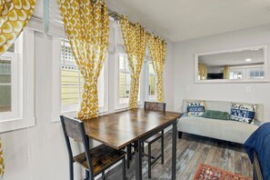 A dining area with a wooden table and chairs, a sofa with decorative pillows, and a window with yellow patterned curtains. - Your Group's Home Base | Downtown | Hot Tub+Games (Watkins Glen)