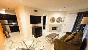 A modern living room with a sofa, a coffee table, and a TV. There is a kitchen area with cabinets and a microwave.