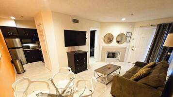 A modern living room with a sofa, a coffee table, and a TV. There is a kitchen area with cabinets and a microwave.