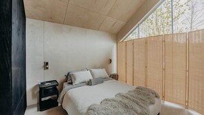 Quarto moderno com cama grande, cabeceira de madeira e vista para árvores pela janela.