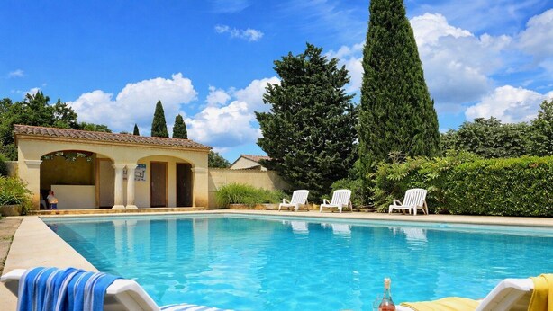 Une piscine avec des chaises longues, un espace abrité avec des colonnes et de grands arbres en arrière-plan.