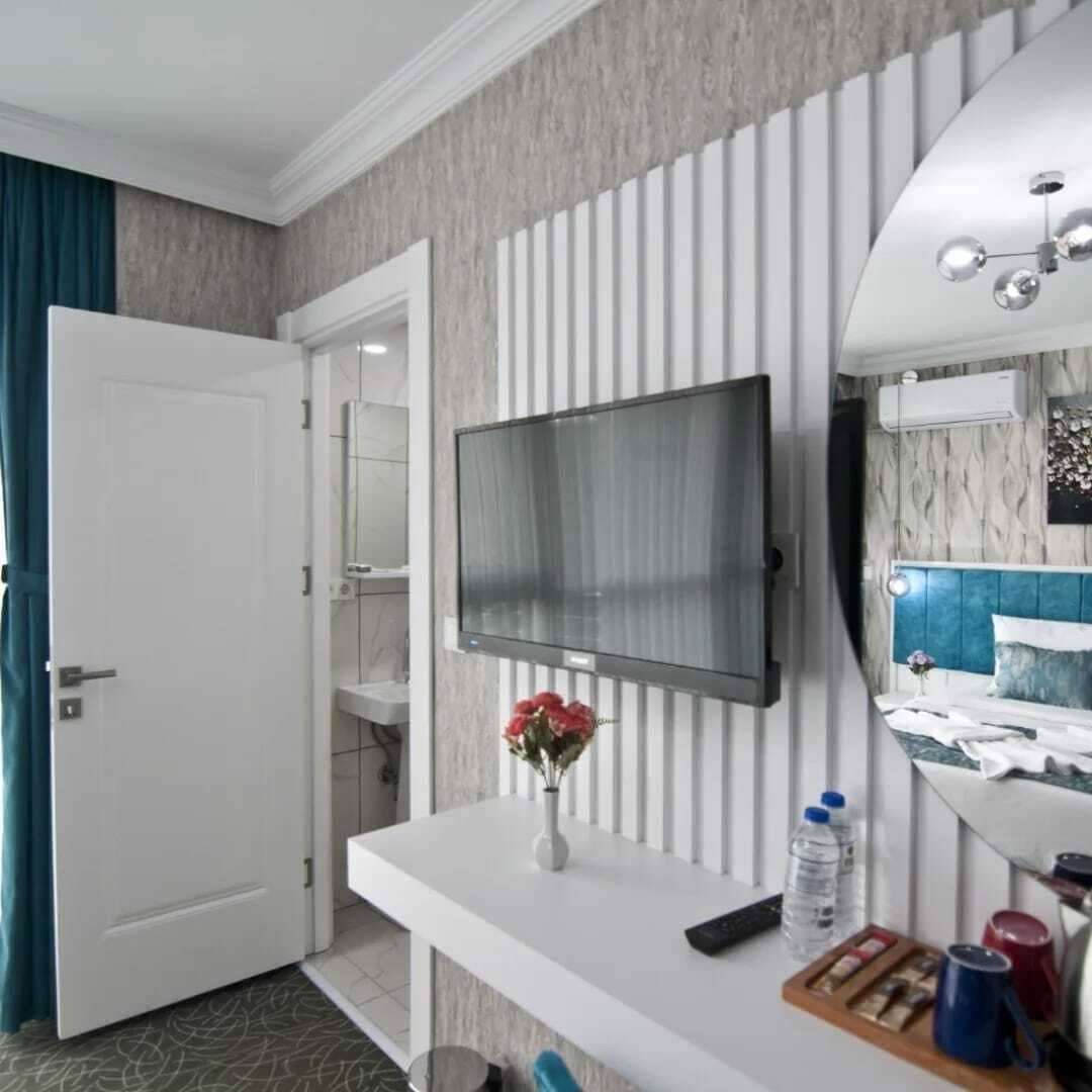 Habitación de hotel con televisor montado en la pared, una mesita consola blanca con un jarrón de flores, cama con cabecera azul y baño con toallero.