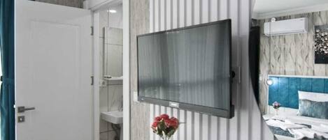 Habitación de hotel con televisor montado en la pared, una mesita consola blanca con un jarrón de flores, cama con cabecera azul y baño con toallero.