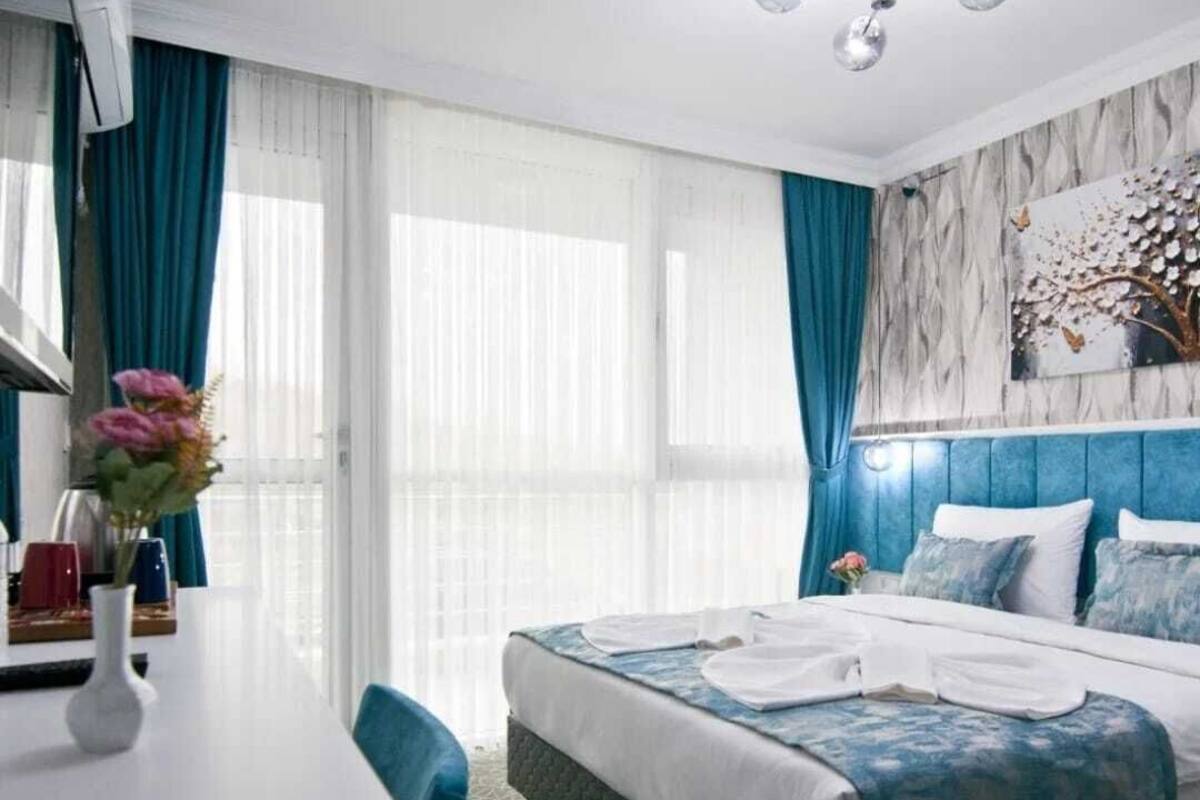A hotel room with a bed, a desk, and a chair. There is a window with curtains, a wall-mounted air conditioner, and a ceiling light fixture.