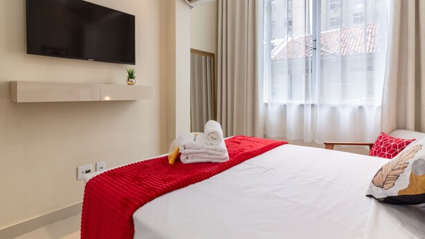 A hotel room with a bed, a TV mounted on the wall, an air conditioning unit, a window with curtains, and a small plant on the bedside table. - Cosy in Copacabana 7 Minutes From the Beach Fm211 (Rio de Janeiro)