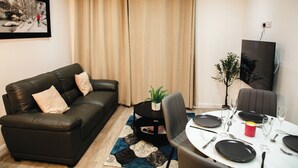 A living room with a black leather sofa, a dining table set for two, and a large window with beige curtains.