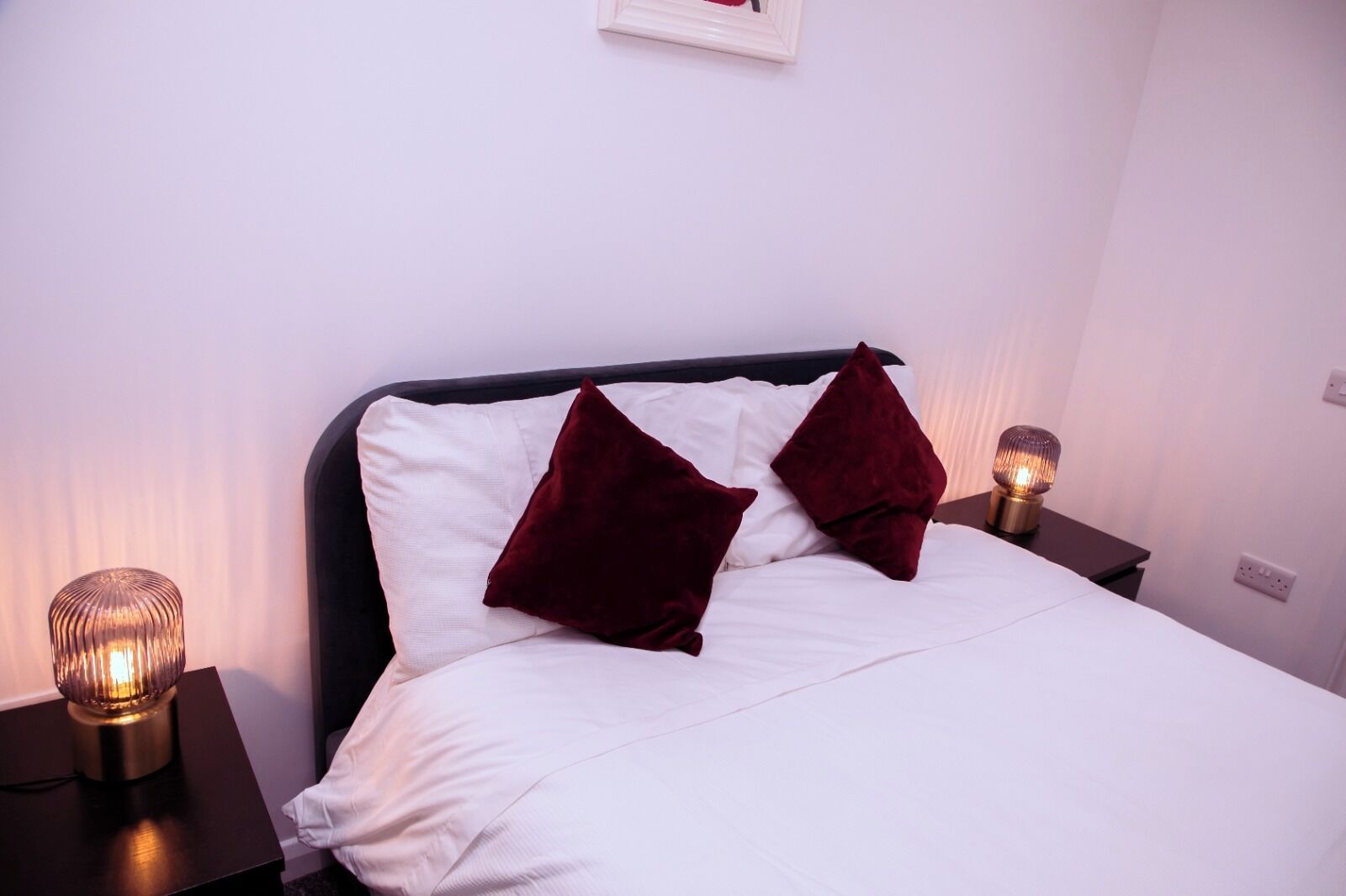 Una cama bien tendida con sábanas blancas y dos almohadas rojo oscuro, flanqueada por dos mesitas de noche con lámparas.