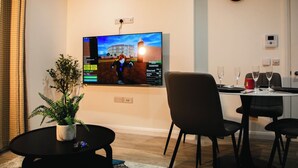 A modern living room with a flat-screen TV mounted on the wall, a dining area with a table and chairs, and a potted plant on a small table.