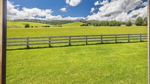 Blick durch ein Fenster auf ein grünes Feld mit Holzzaun, blauem Himmel und ferne Berge.