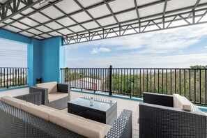 Un espacio al aire libre techado con mobiliario de mimbre y una mesa de vidrio, con vistas a un jardín ajardinado.