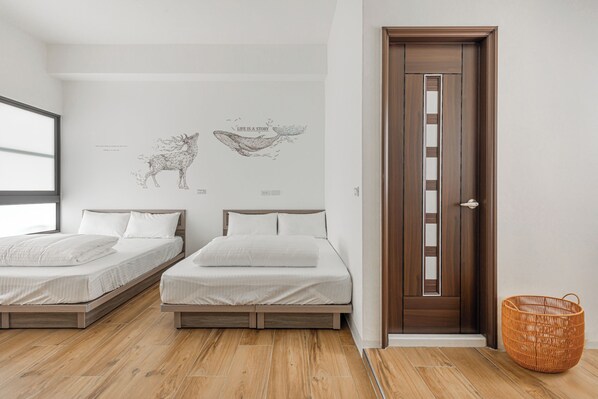 Un dormitorio con dos camas individuales, suelo de madera, un ventanal grande y una puerta con una cesta a un lado.