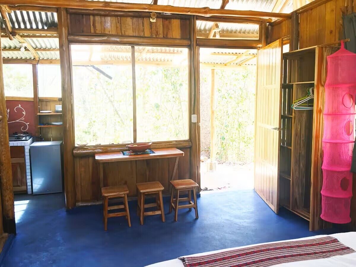 A wooden cabin with a kitchen area, a dining table with stools, and a large window.
