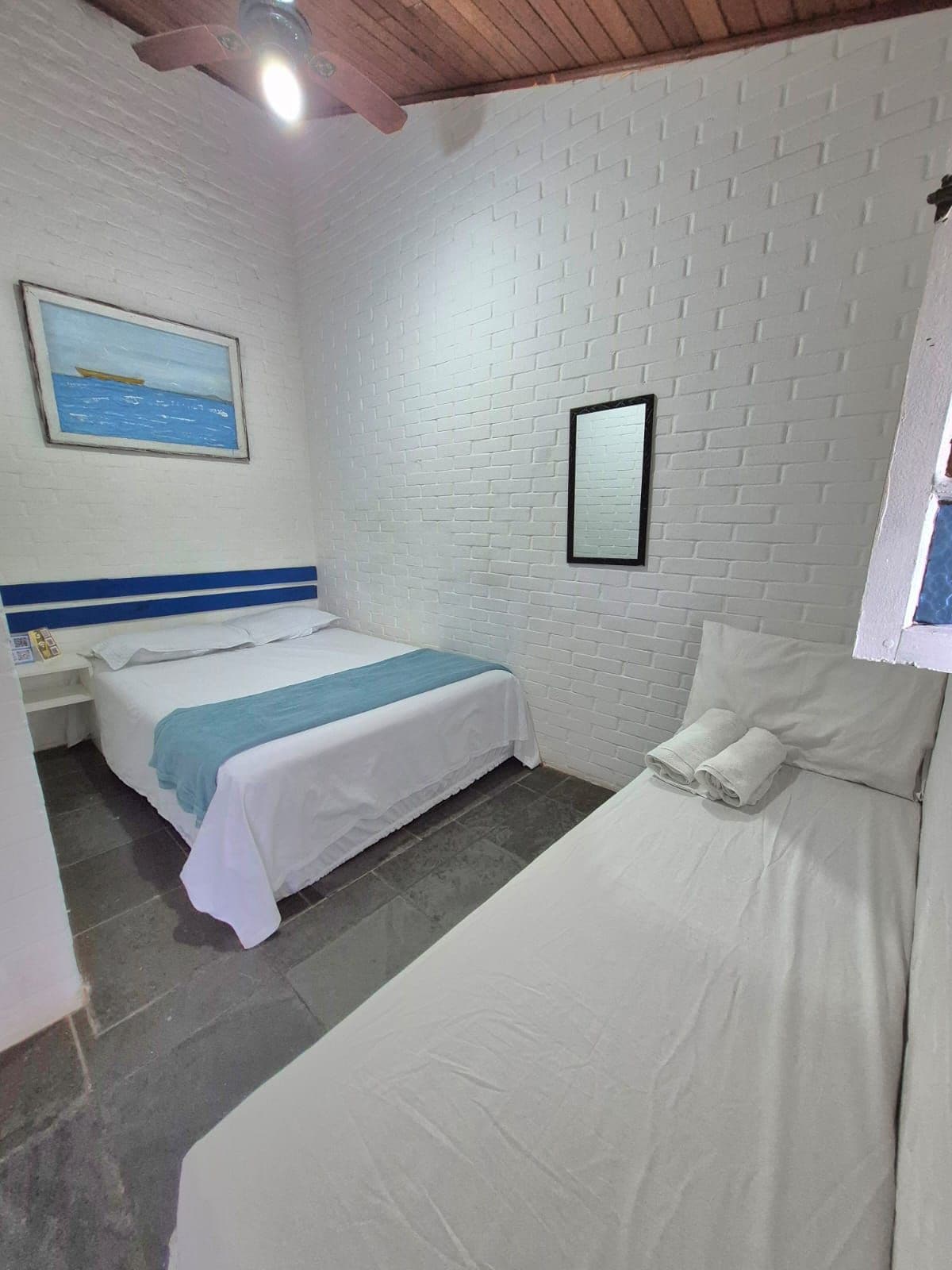 Una habitación con dos camas, una pared de ladrillo blanco, un ventilador de techo y un cuadro de un barco en la pared.