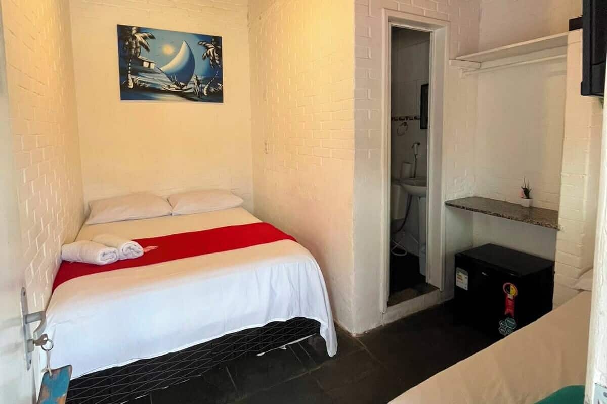 Una habitación con una cama, un ventilador de techo, un cuadro, una puerta y un estante pequeño.