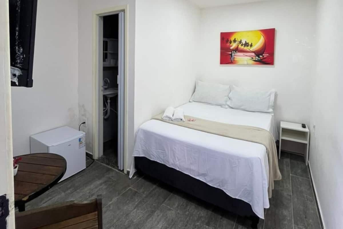 Un dormitorio con cama, escritorio, silla, un cuadro en la pared y una puerta que da al baño.