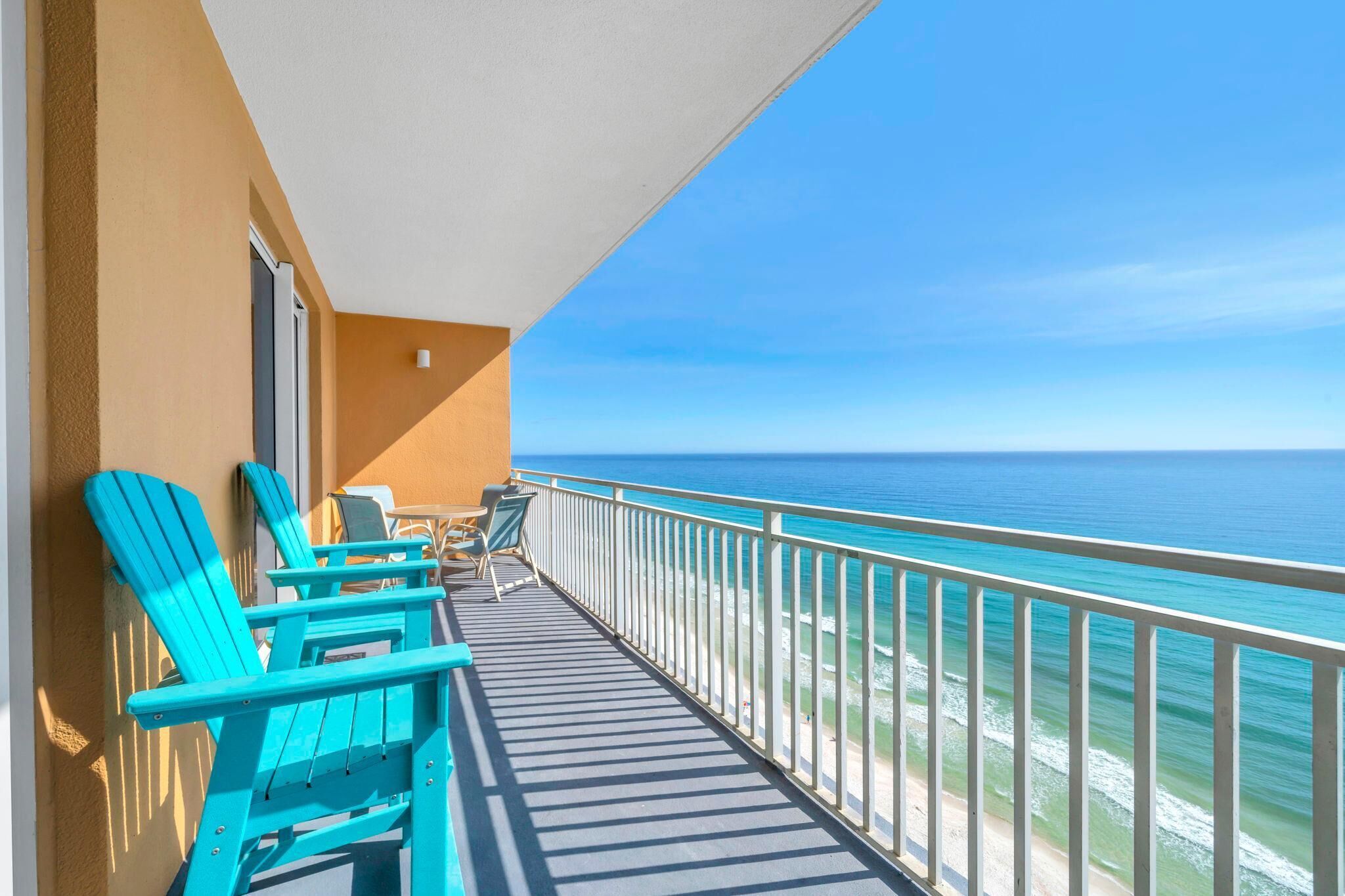 A balcony with turquoise chairs and a table, overlooking a beach with clear blue water.