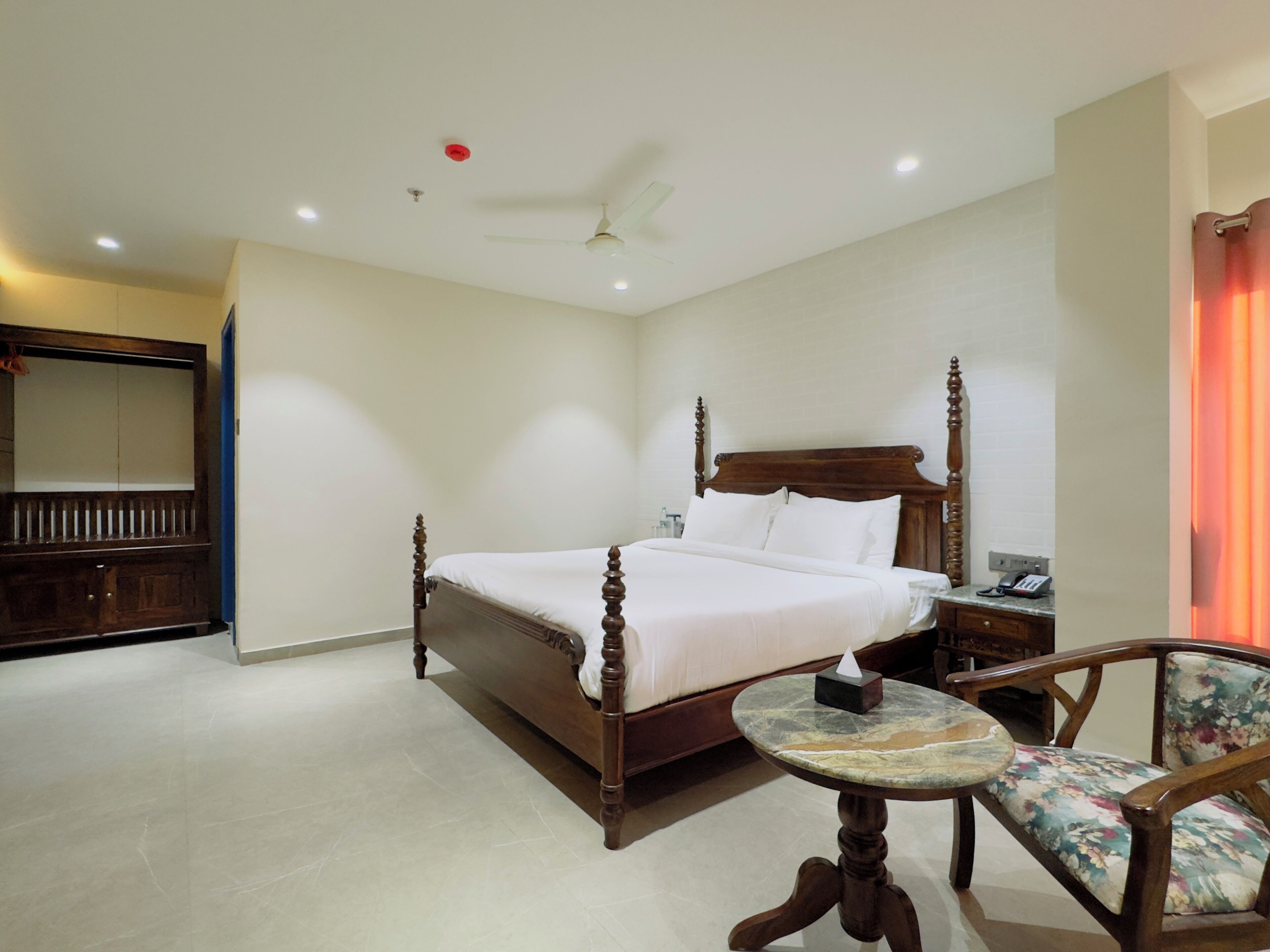 A hotel room with a wooden bed, a small round table, a chair, a mirror, and a ceiling fan.