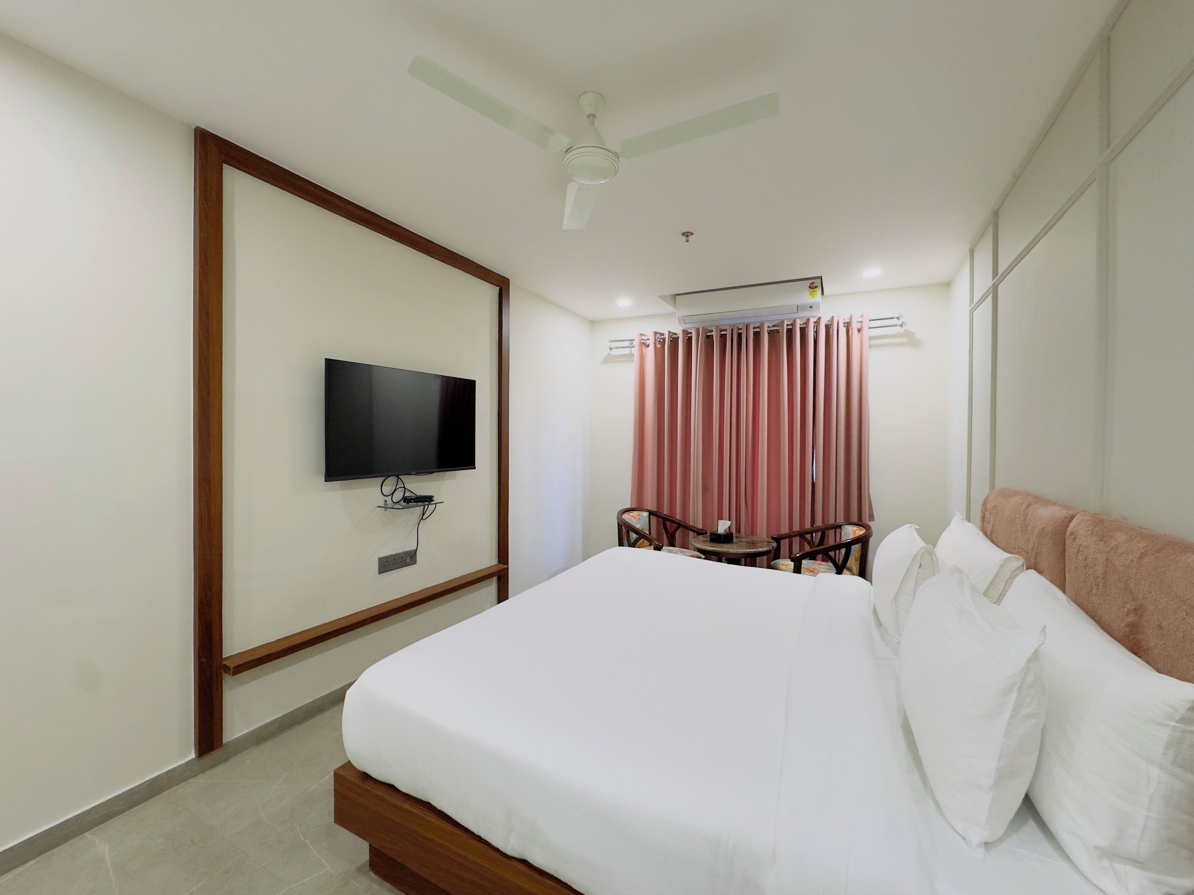 A hotel room with a bed, a television mounted on the wall, a ceiling fan, and a window with curtains.