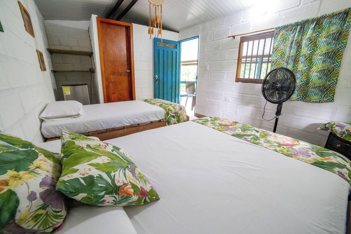 Quarto com duas camas, uma porta azul, um ventilador e uma janela com grades.