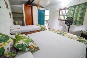 Una habitación con dos camas, una puerta azul, un ventilador y una ventana con barrotes.