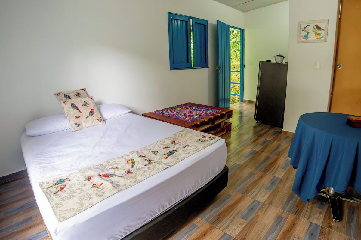 Quarto com cama, um banquinho de madeira, mesa e geladeira.
