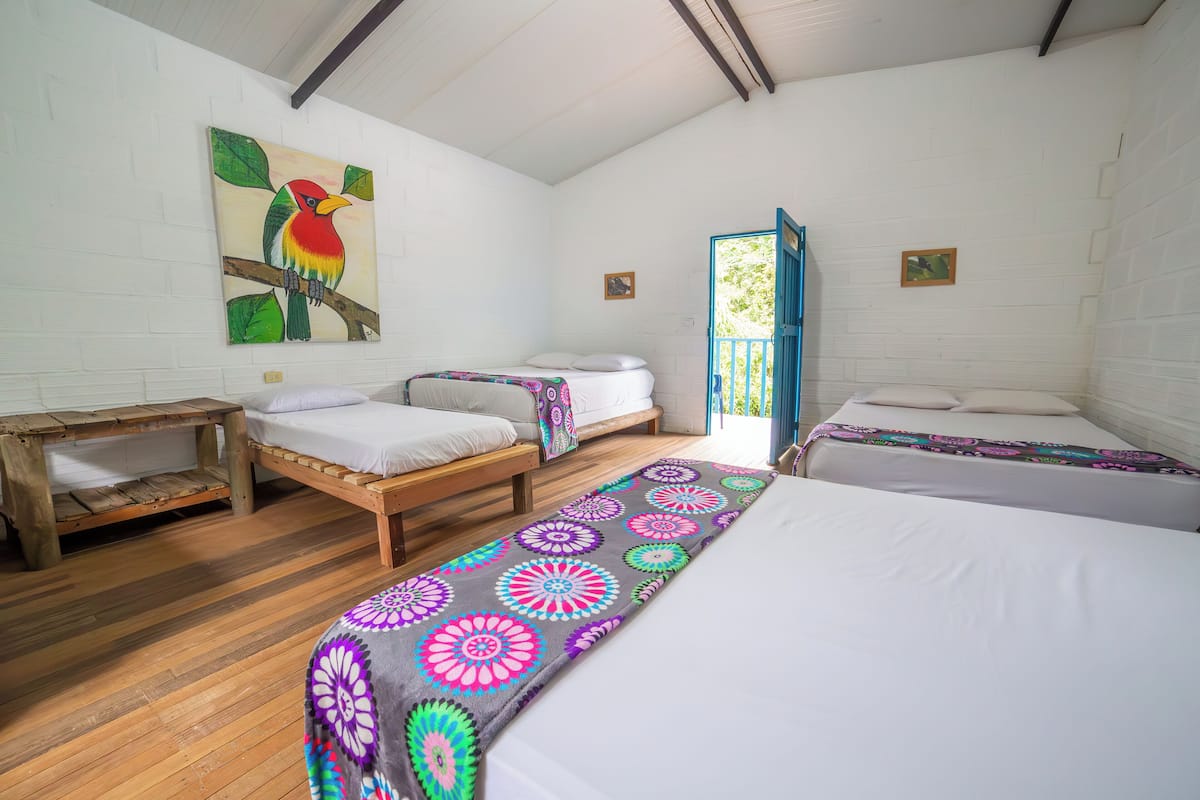 Quarto com duas camas de solteiro, um banco de madeira, um tapete colorido e um quadro de um pássaro na parede.