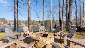 A deck with Adirondack chairs, a stone fire pit, and a view of a lake and trees.