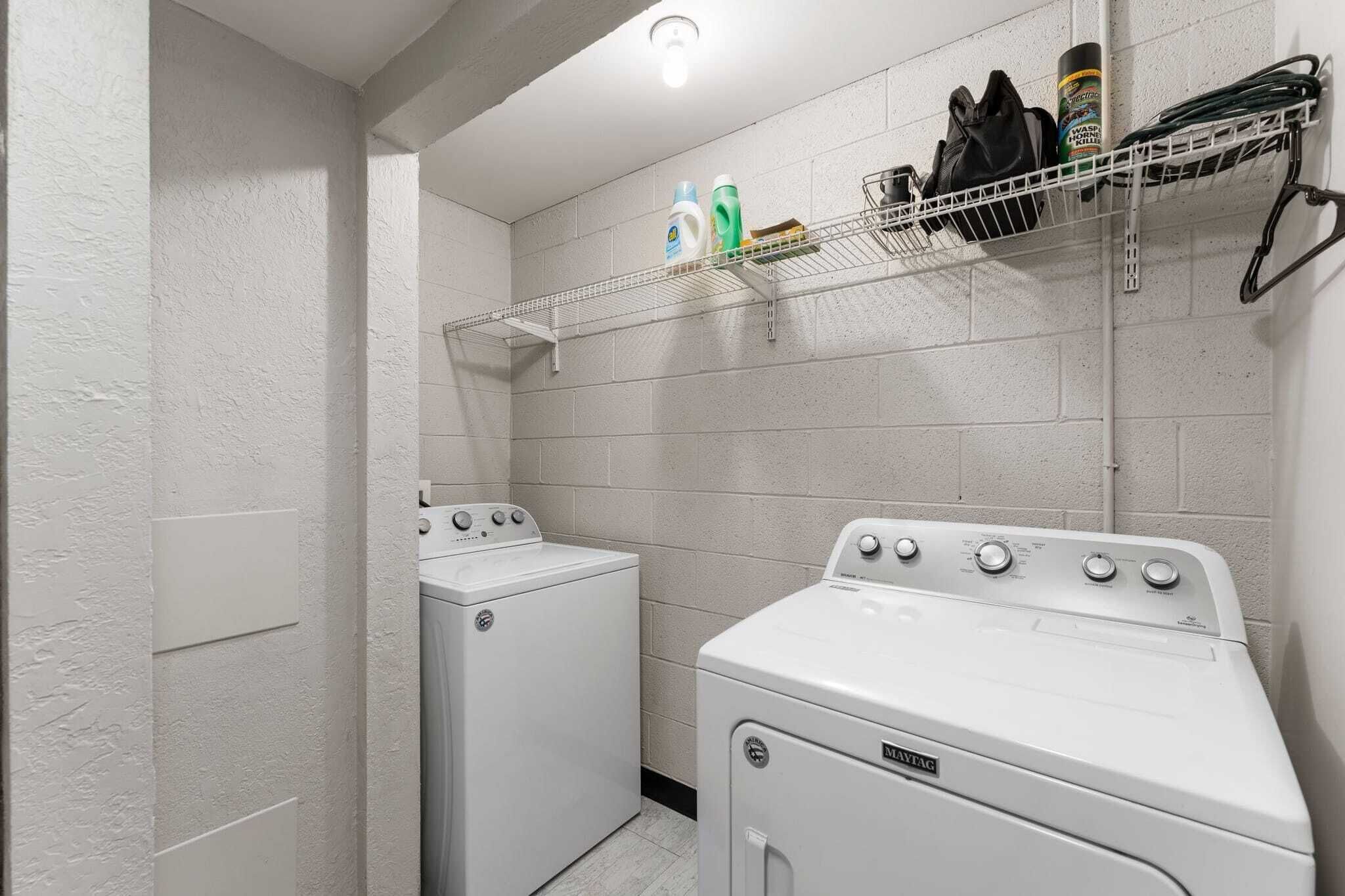 A laundry room with two white front-loading washers and dryers, a metal shelf with cleaning supplies, and a white brick wall.