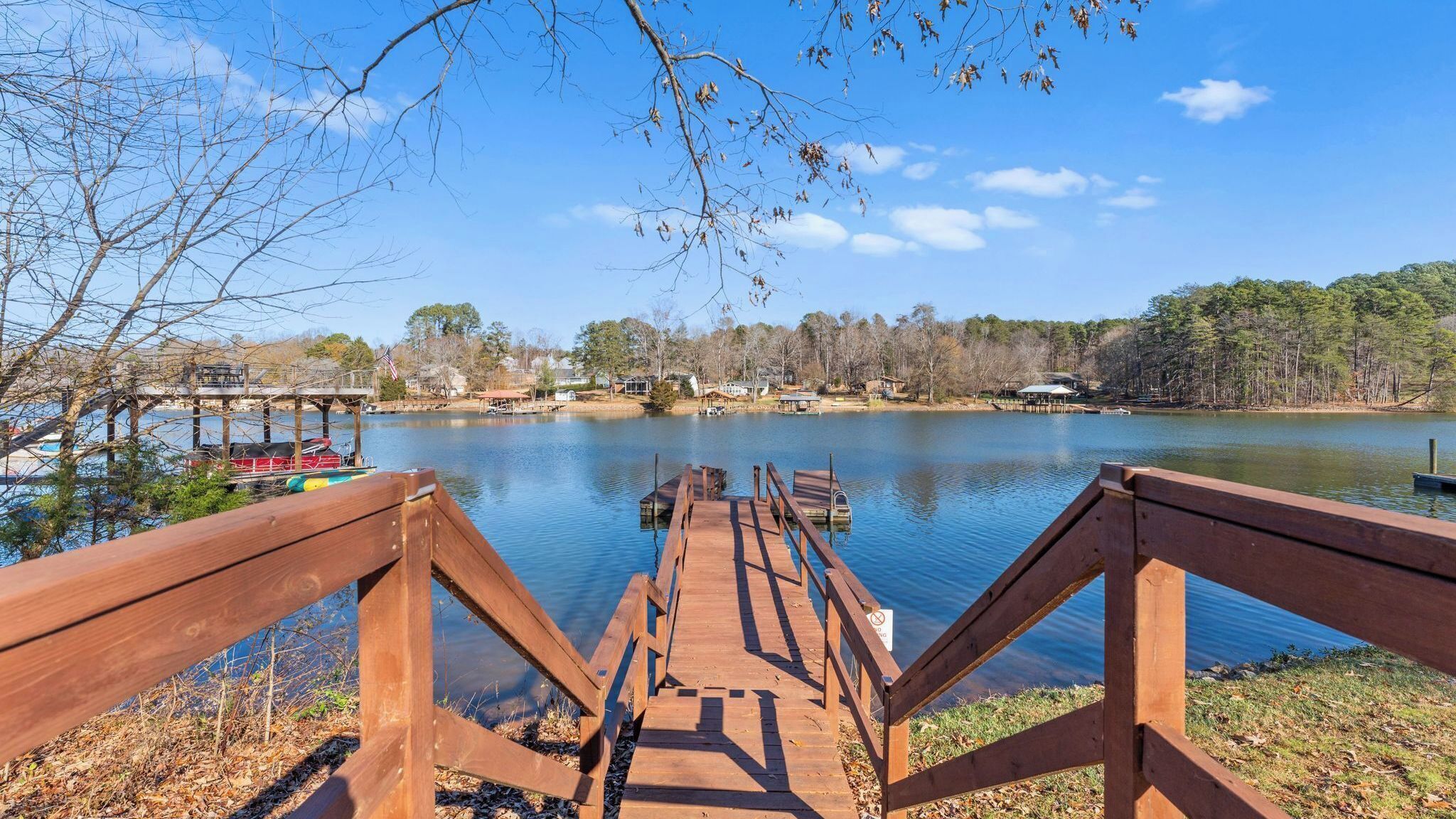 A wooden dock leading to a calm lake with boats, surrounded by trees and houses.
