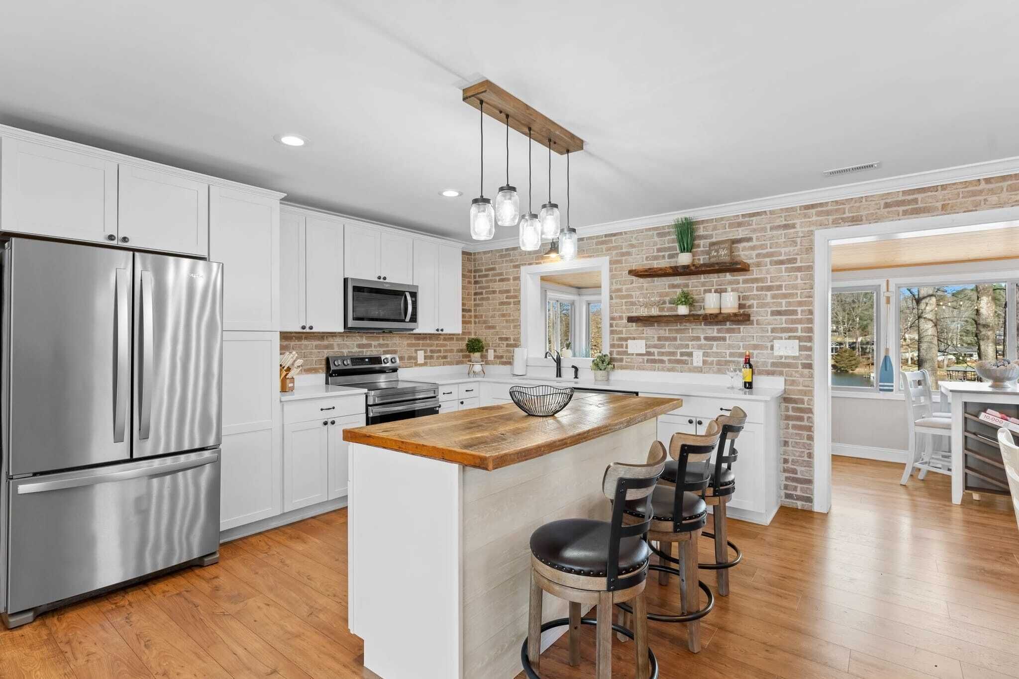A modern kitchen with white cabinets, a stainless steel refrigerator, a wooden island with bar stools, and a brick accent wall.