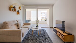 Un salon moderne avec un canapé, un téléviseur posé sur un meuble en bois, une petite table avec une plante, et une vue sur le balcon extérieur.
