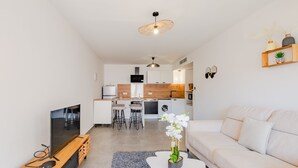 Un salon moderne avec un canapé, une table basse et un téléviseur. Au fond, on aperçoit une cuisine avec un coin repas.