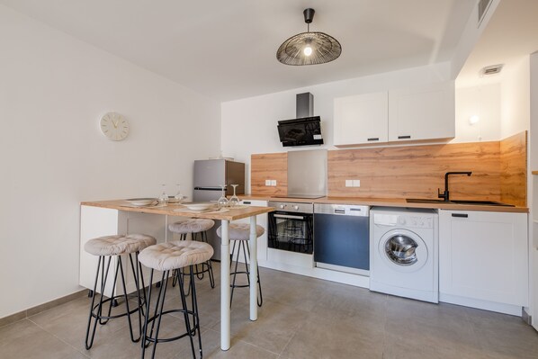 Una cocina compacta con encimera de madera, armarios blancos, horno integrado, lavavajillas y lavadora. Hay taburetes de bar y una lámpara colgante.
