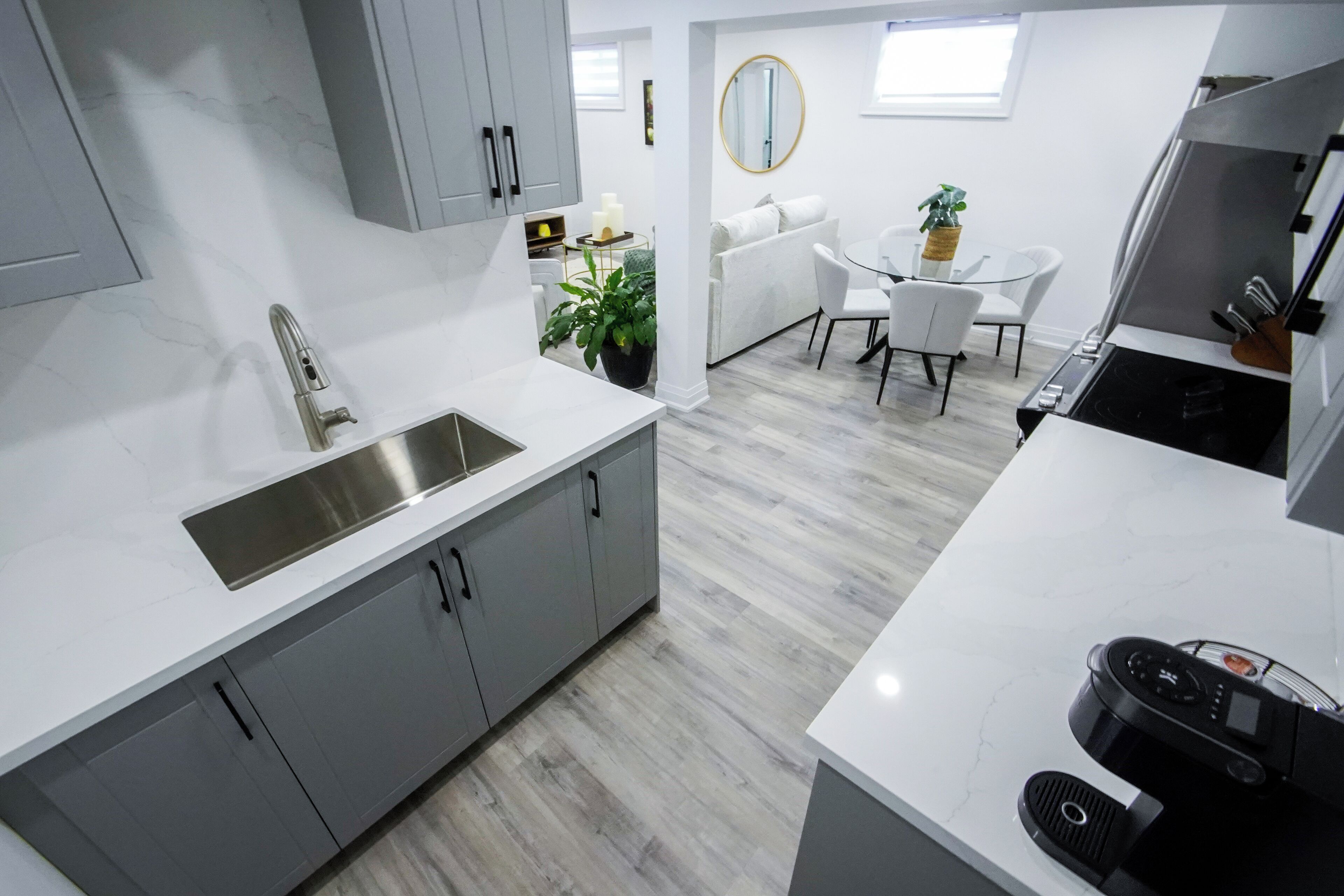 A modern kitchen with grey cabinets, a stainless steel sink, and a white countertop. There is a dining area with a white table and chairs, and a small potted plant.