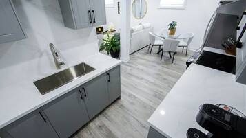 A modern kitchen with grey cabinets, a stainless steel sink, and a white countertop. There is a dining area with a white table and chairs, and a small potted plant.