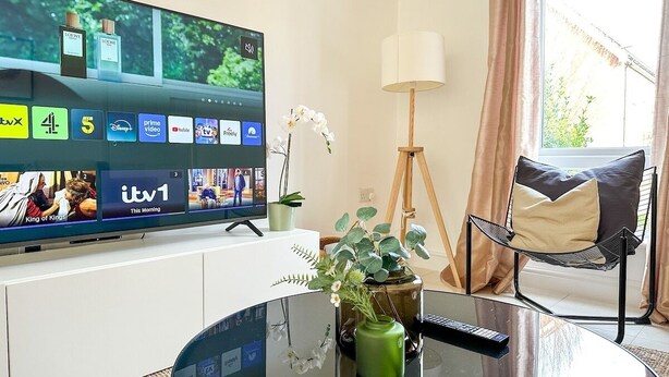 A modern hotel room with a large flat-screen TV displaying various channels, a round glass coffee table with a vase of flowers, a comfortable chair, and a floor lamp.