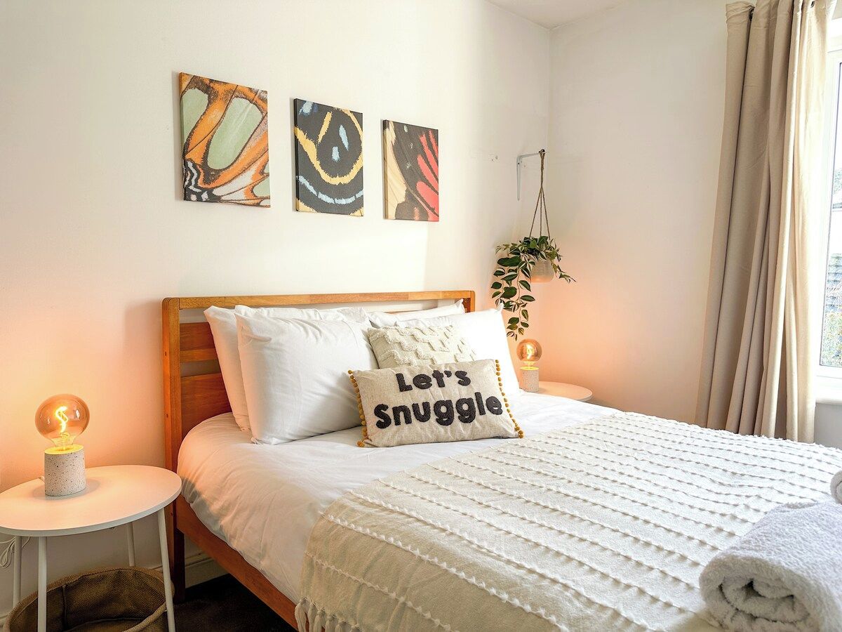 A neatly made bed with a pillow that reads 'Let's Snuggle,' a bedside lamp, and wall art above the bed.