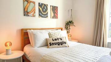 A neatly made bed with a pillow that reads 'Let's Snuggle,' a bedside lamp, and wall art above the bed.