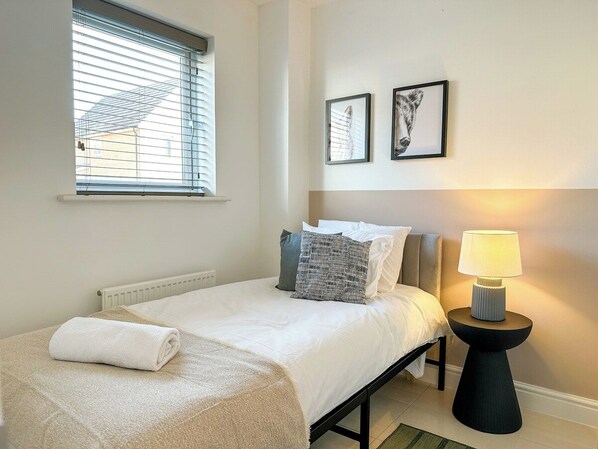 A bedroom with a bed, a bedside lamp, a window with blinds, and two framed pictures on the wall.
