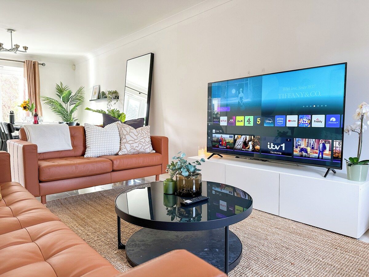 A modern living room with a leather sofa, a glass coffee table, and a flat-screen TV displaying various channels.