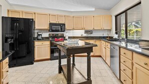 A modern kitchen with wooden cabinets, a black refrigerator, a microwave, a coffee maker, and a large window providing a view of greenery outside. - Riverside Retreat: 4BR/3BA with Private Dock and Boat Lift (Stuart)