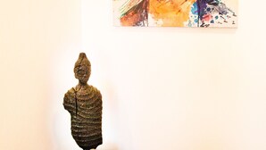 A painting of a person with a floral headpiece on a wall, a sculpture on a wooden table, and a radiator below.