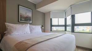En snyggt bäddad säng med vita sängkläder och en beige pläd, en inramad bild av en stadsbild på väggen och ett fönster med persienner.
