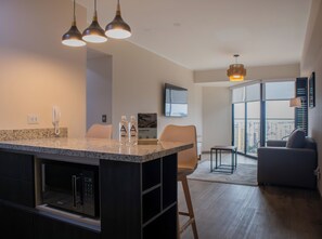 Een moderne keuken met granieten aanrecht, een ingebouwde oven en een koffieapparaat. Er is een zithoek met een tafel en stoelen, een bank en een groot raam met uitzicht op de stad.