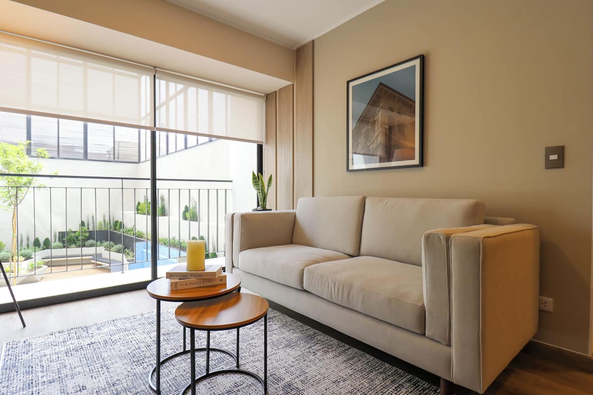 Ein Wohnzimmer mit einer Couch, einem kleinen runden Tisch mit einem Buch und einem großen Fenster mit Blick auf einen Garten und ein Gebäude.
