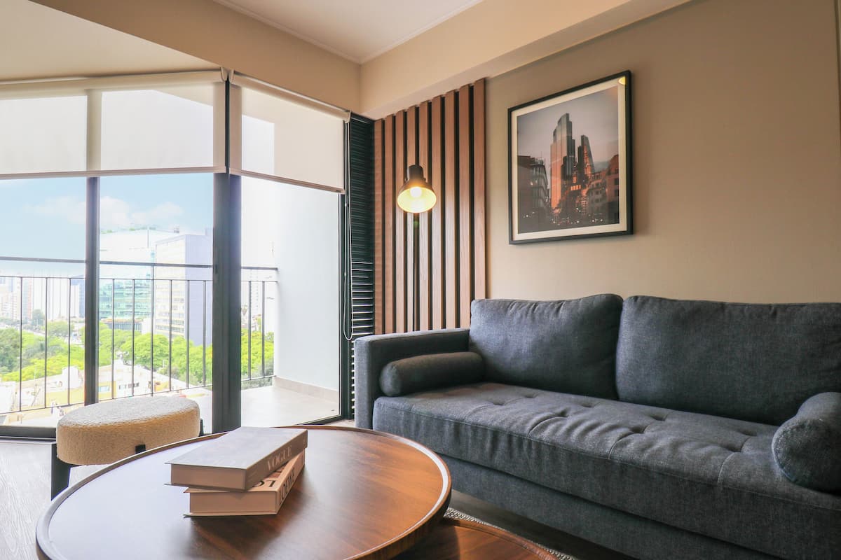 Ein modernes Wohnzimmer mit einem grauen Sofa, einem runden Couchtisch mit Büchern, einem Stadtbild als gerahmtes Bild an der Wand und einem großen Fenster mit Jalousien.