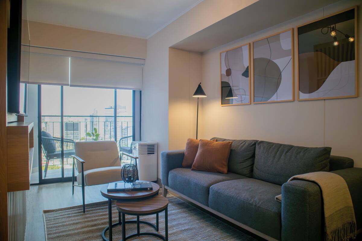 Ein modernes Wohnzimmer mit einer Couch, einem Sessel und einem Couchtisch. Ein großes Fenster bietet Blick auf Gebäude.