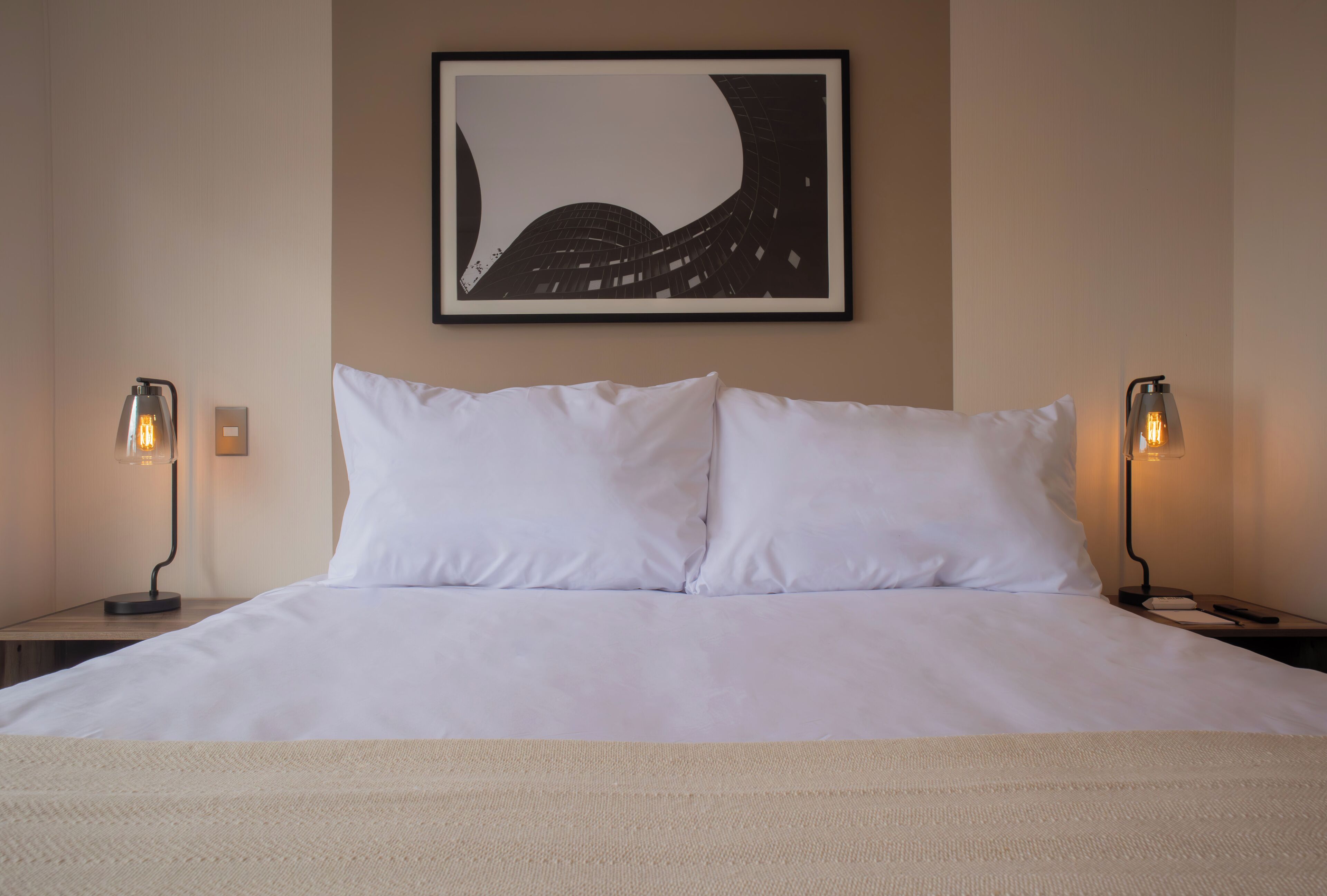 Un lit bien fait, avec du linge de lit blanc, deux lampes de chevet et une œuvre d’art encadrée au mur.