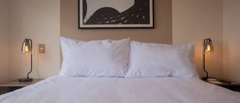 Un lit bien fait, avec du linge de lit blanc, deux lampes de chevet et une œuvre d’art encadrée au mur.