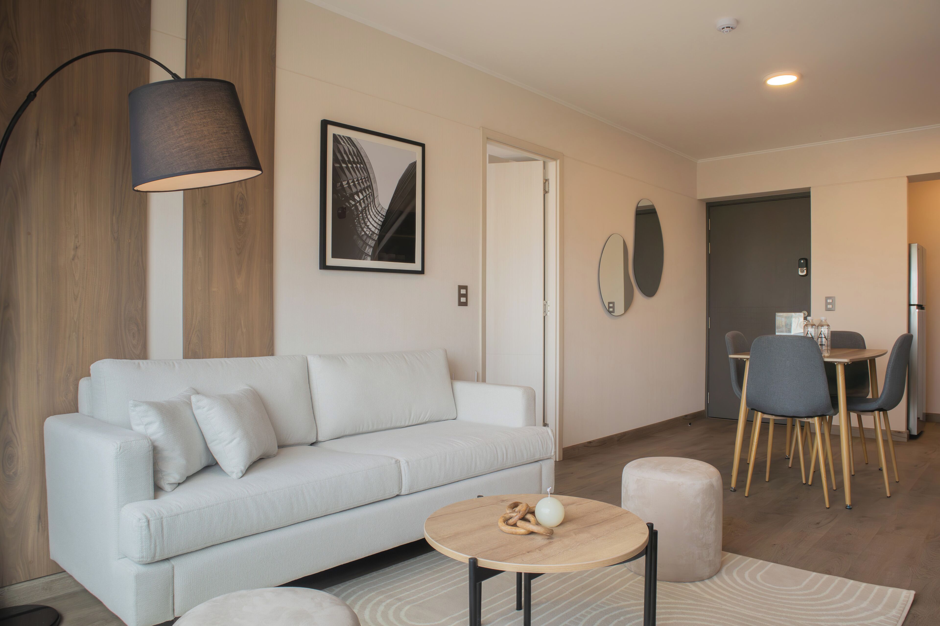 Una sala de estar moderna con un sofá blanco, una mesa de centro redonda de madera y un comedor con una pequeña mesa y sillas.